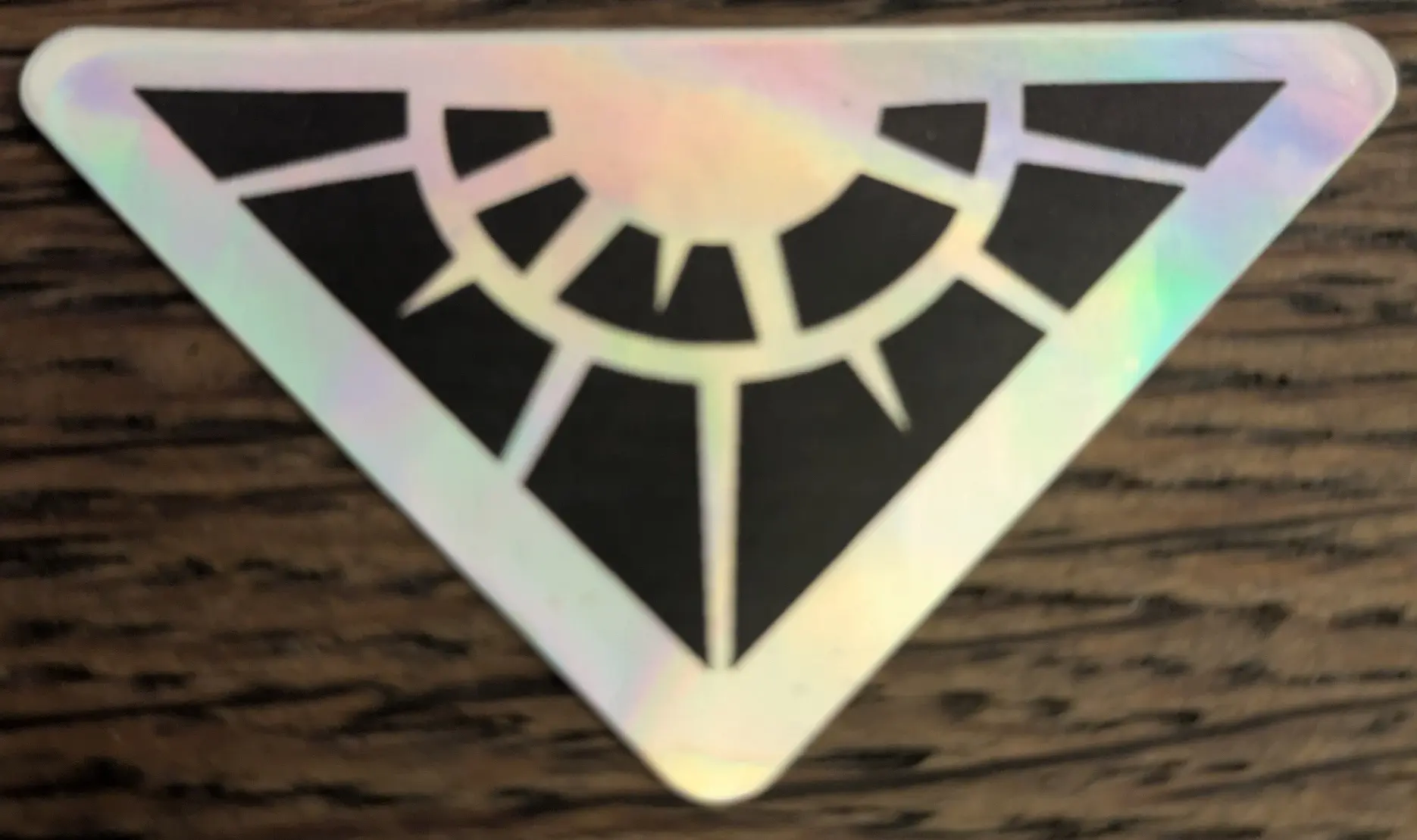 A silvery sticker (shining in many colours like a puddle of oil) of the Spellbreak video game logo on a somewhat black wooden desk.